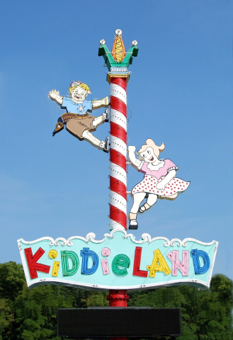 The sign that originally stood outside of the Kiddieland park.  "Kiddieland" is written in bright, multicolored letters, mounted on a red and white striped pole alongside two happy children who seem excited to enjoy the park.