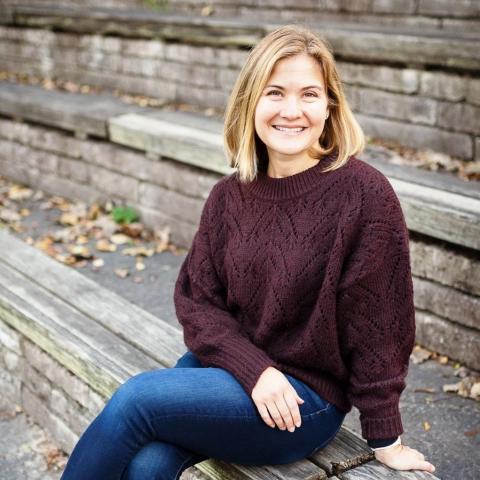 A portrait of Leah Bostrom, a blonde woman wearing an oversized maroon sweater and jeans sits on a low brick wall.