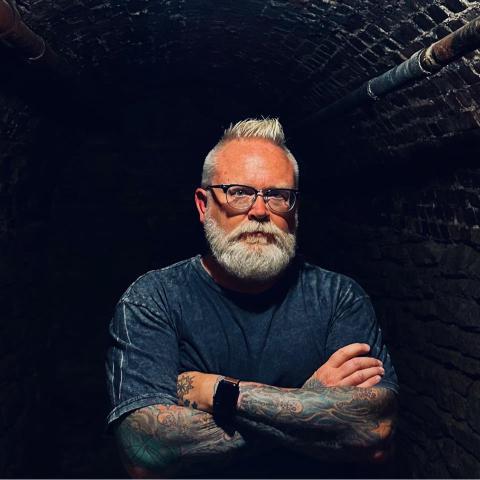 A white haired bearded man in glasses and a blue tshirt standing against a dark background.