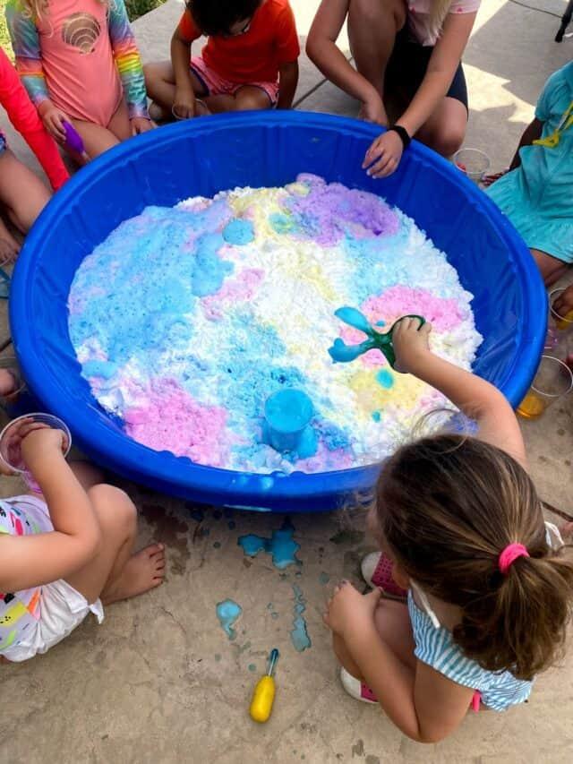 Kiddie pool filled with baking soda and surrounded by children. The children are adding colorful vinegar to the baking soda. 