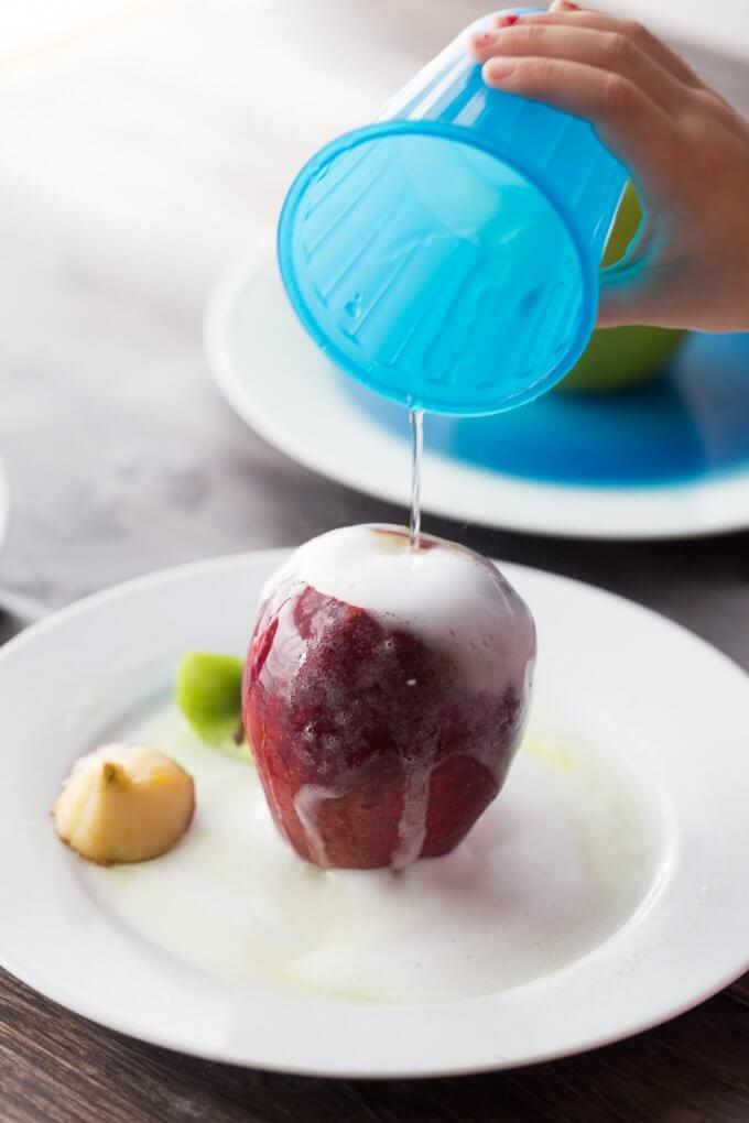 hand holding a blue cup with liquid being poured over an apple with foam coming out of apple.