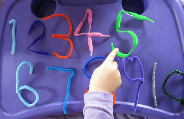 Various colors of fuzzy pipe cleaners in the shape of numbers with a small hand pointing towards the number five.