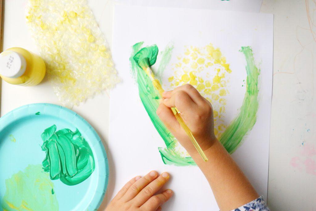 toddler painting green leaf next to bubble wrap-stamped corn 